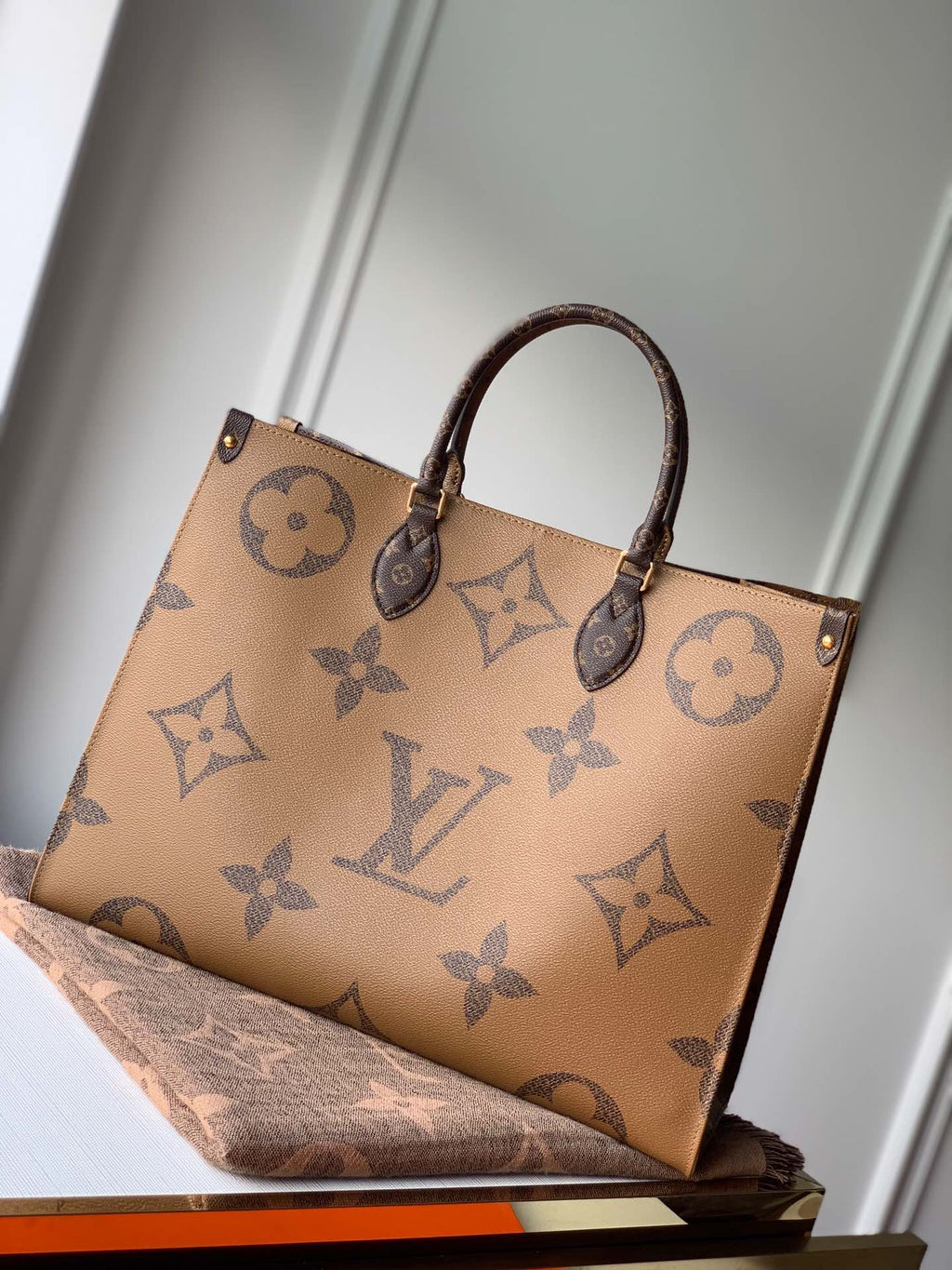 Tan tote bag with oversized dark brown monogram motifs, double top handles, and gold-tone hardware.